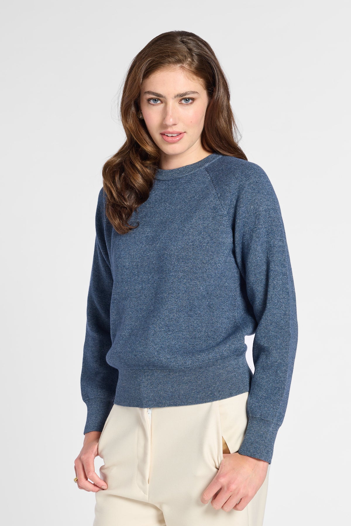 A woman modeling a heathered slate blue knit sweater with raglan sleeves, paired with cream-colored trousers, against a plain light gray studio background.