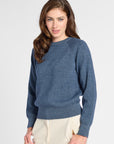 A woman modeling a heathered slate blue knit sweater with raglan sleeves, paired with cream-colored trousers, against a plain light gray studio background.