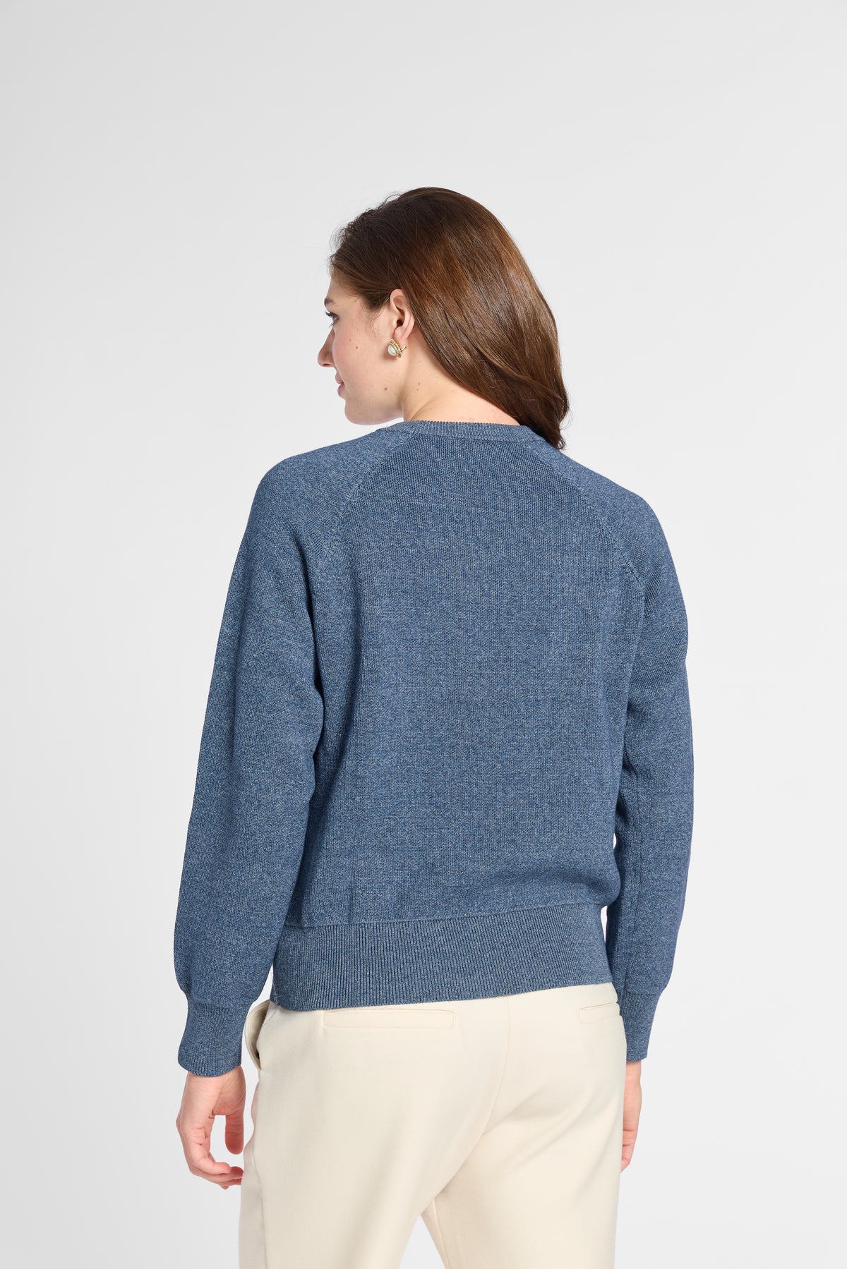A woman viewed from the back wearing a heathered blue crewneck sweater and cream-colored pants, set against a plain light gray studio background.