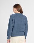 A woman viewed from the back wearing a heathered blue crewneck sweater and cream-colored pants, set against a plain light gray studio background.