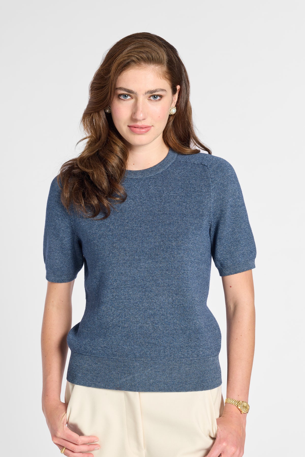 A woman modeling a short-sleeve crewneck knit sweater in a heathered slate blue color, paired with cream-colored trousers, against a plain light gray studio background.