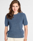 A woman modeling a short-sleeve crewneck knit sweater in a heathered slate blue color, paired with cream-colored trousers, against a plain light gray studio background.