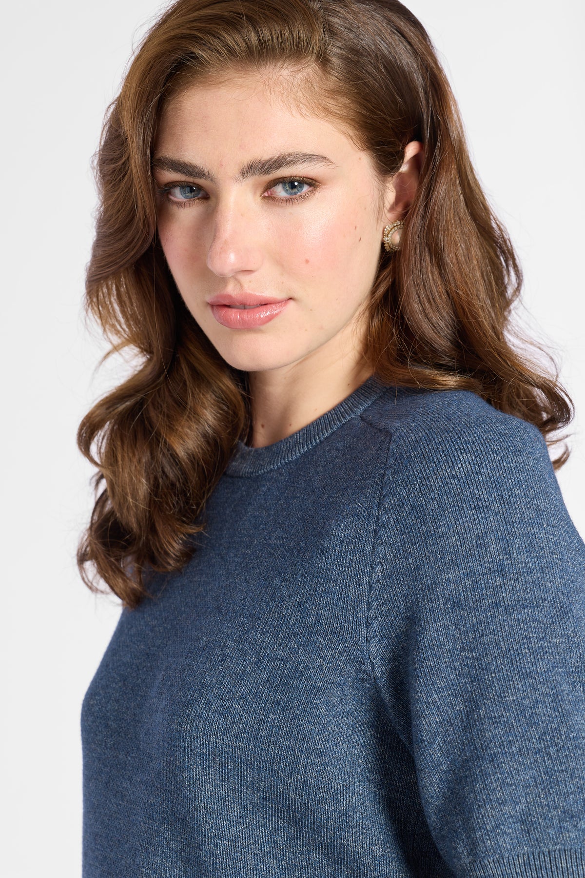 A woman with wavy brown hair wearing a textured slate blue knit sweater, shown in a close-up portrait against a light gray background.