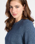 A woman with wavy brown hair wearing a textured slate blue knit sweater, shown in a close-up portrait against a light gray background.