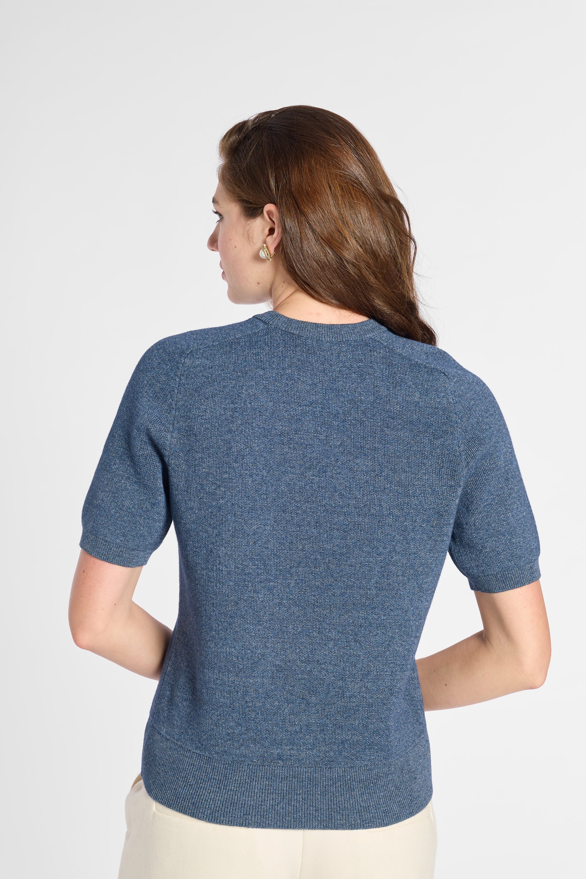 A woman shown from the back wearing a short-sleeve textured knit top in heathered blue, paired with light beige pants, against a plain light gray studio background.