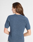 A woman shown from the back wearing a short-sleeve textured knit top in heathered blue, paired with light beige pants, against a plain light gray studio background.