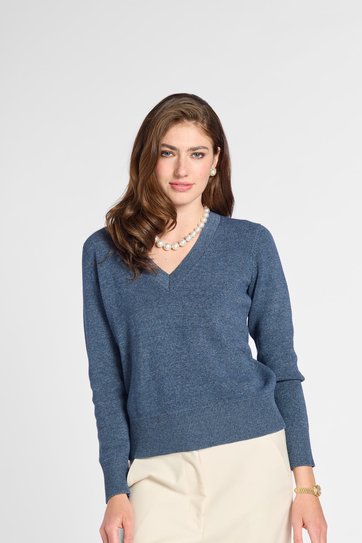 A woman modeling a heathered slate blue V-neck knit sweater with long sleeves, paired with cream pants and a pearl necklace, against a light gray studio background.