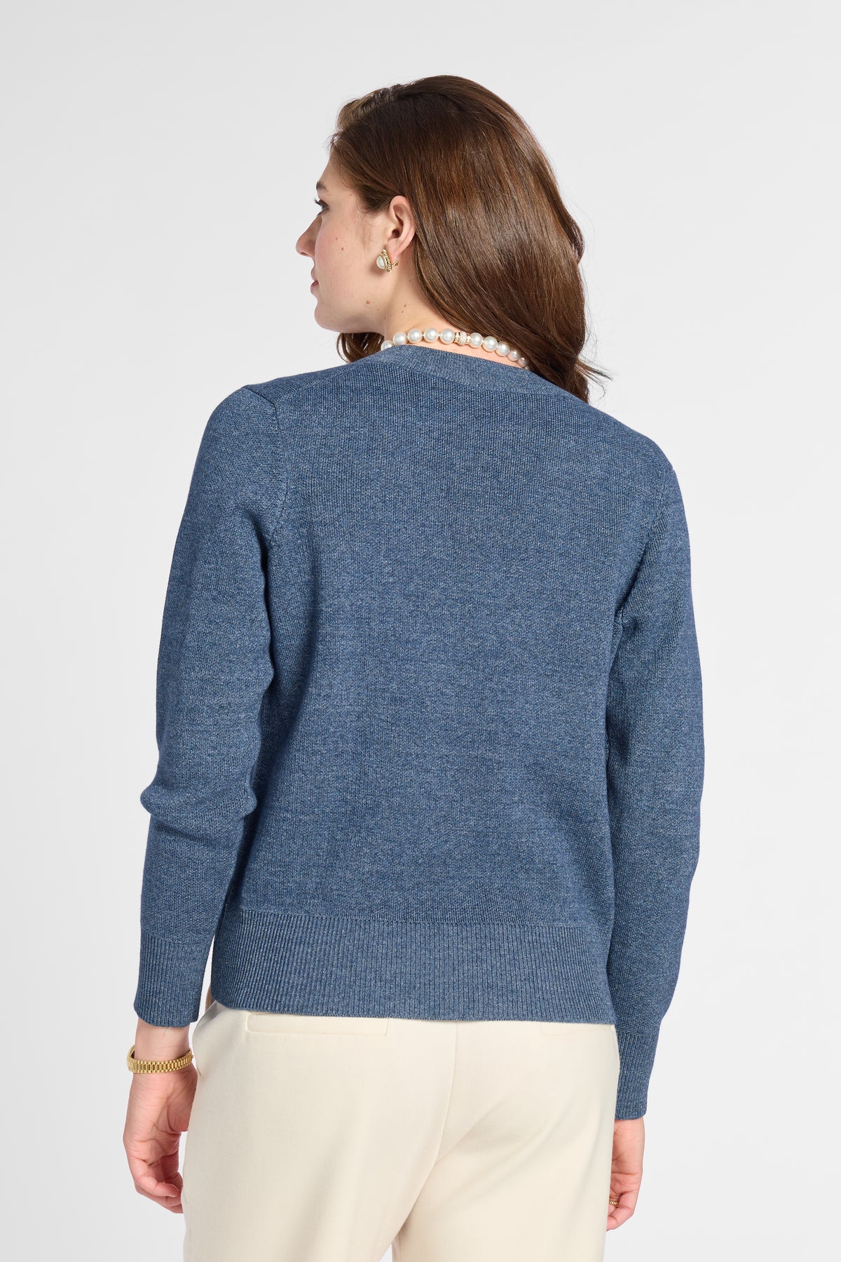 A woman shown from the back wearing a heathered blue knit sweater and cream-colored trousers, photographed against a plain light gray studio background.
