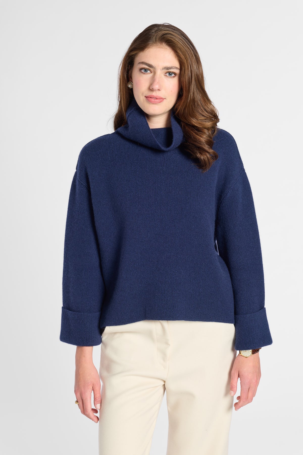 A woman wearing a navy blue knit turtleneck sweater with rolled cuffs and cream-colored trousers, standing against a light gray studio background.