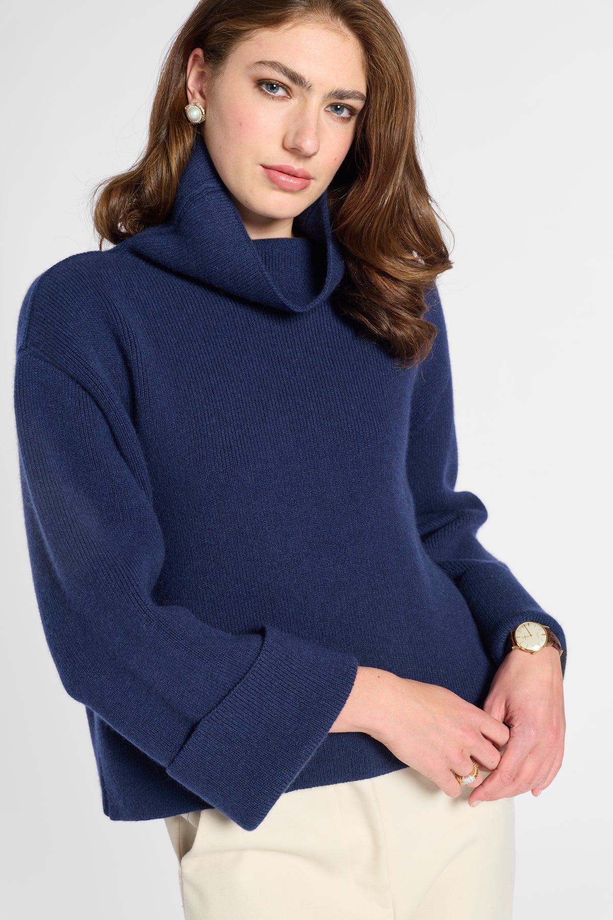A woman wearing a navy blue ribbed knit turtleneck sweater with wide three-quarter sleeves, paired with cream trousers, against a light gray studio background.