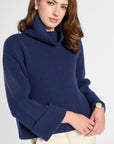 A woman wearing a navy blue ribbed knit turtleneck sweater with wide three-quarter sleeves, paired with cream trousers, against a light gray studio background.