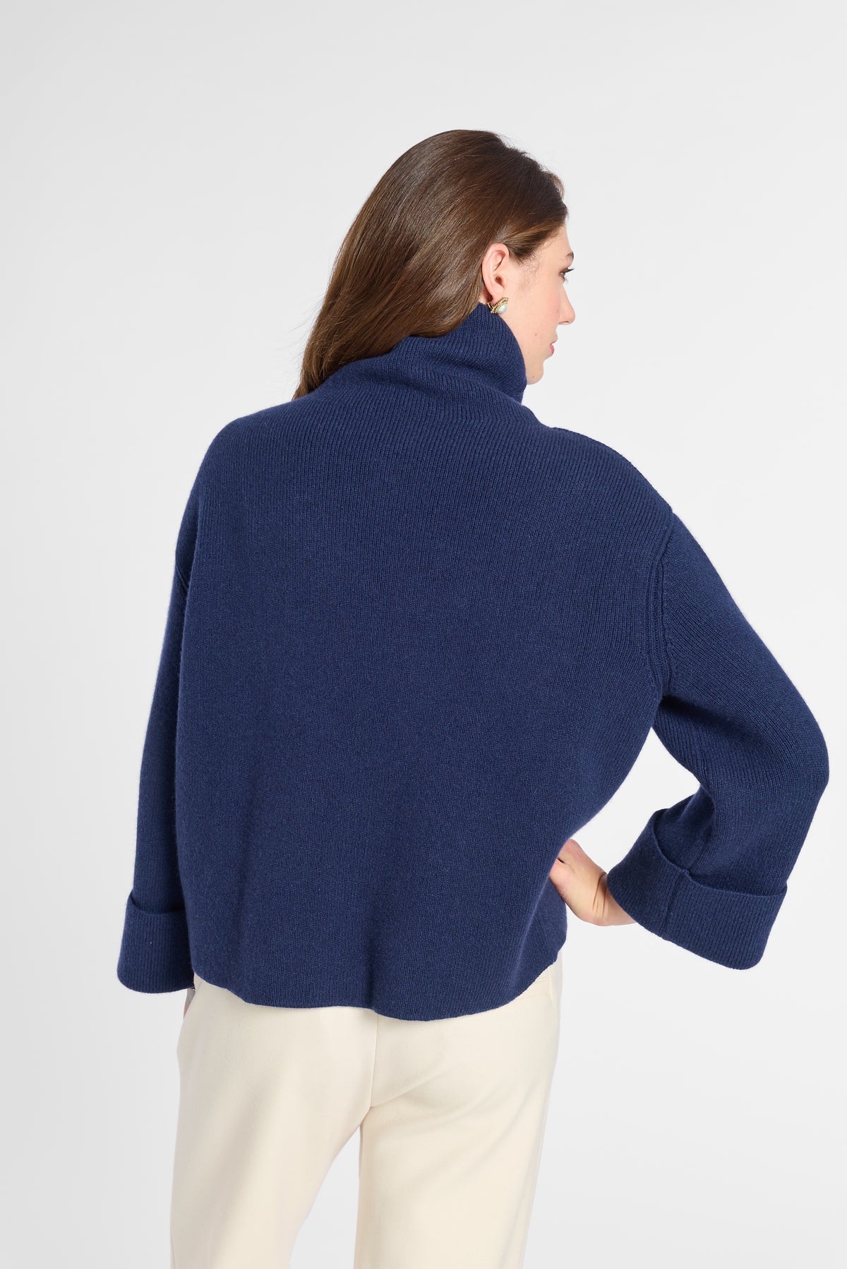 A woman shown from the back wearing a navy blue cropped turtleneck sweater and cream-colored trousers, photographed against a clean white studio background.