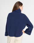 A woman shown from the back wearing a navy blue cropped turtleneck sweater and cream-colored trousers, photographed against a clean white studio background.