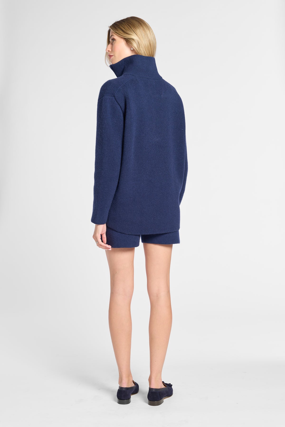 A woman shown from the back wearing a navy blue mock-neck sweater and matching navy shorts against a light gray studio background.