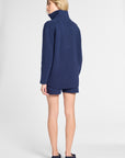 A woman shown from the back wearing a navy blue mock-neck sweater and matching navy shorts against a light gray studio background.