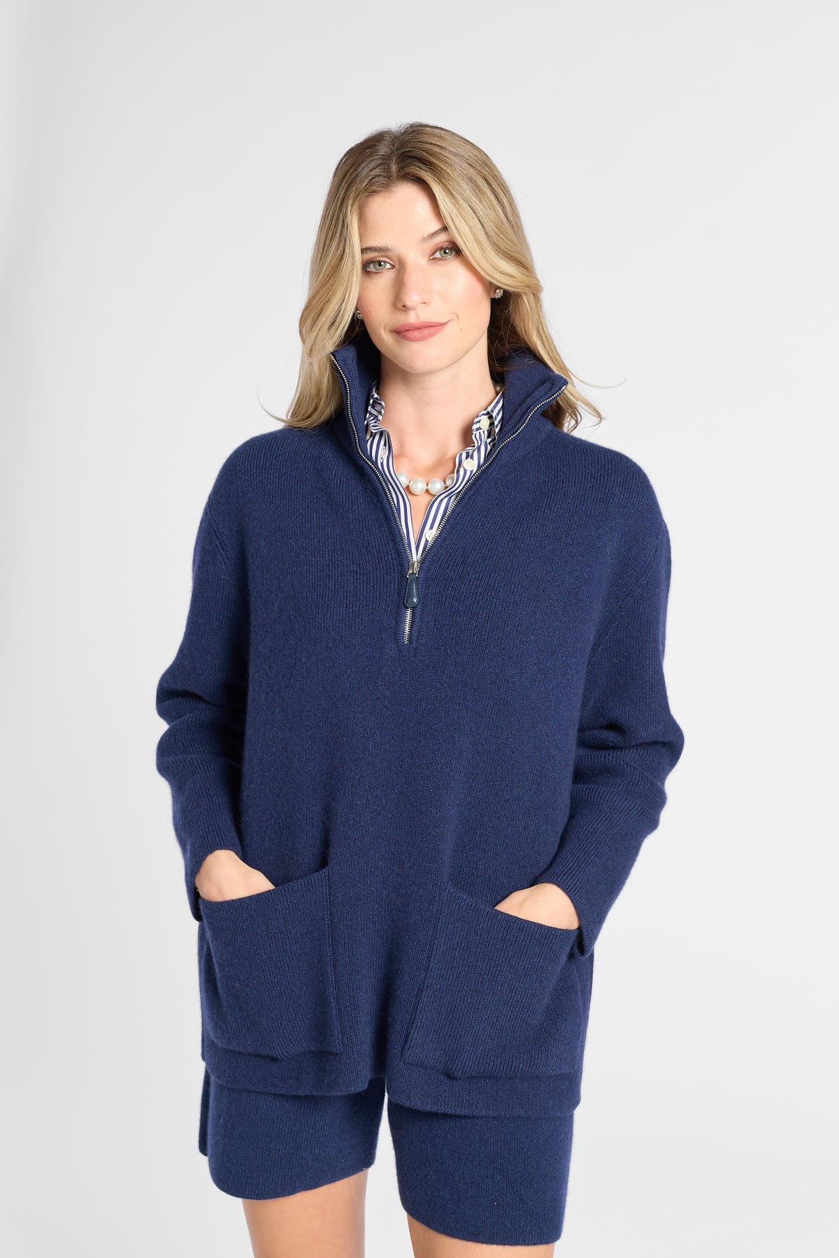 A woman modeling a navy blue knit quarter-zip sweater with matching shorts, featuring a striped inner collar and large front patch pockets, set against a plain light gray studio background.