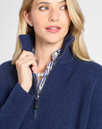 A woman wearing a navy blue ribbed knit quarter-zip sweater over a blue and white striped collared shirt, shown in a close-up shot against a light gray background.