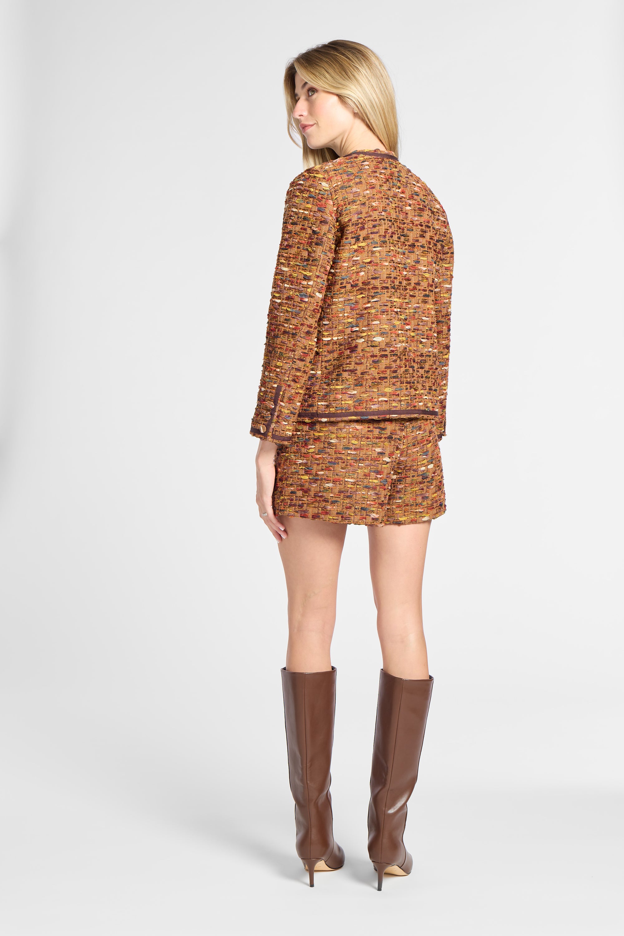 A woman shown from the back wearing a matching tweed jacket and mini skirt set in a warm orange-brown pattern, paired with brown knee-high boots, against a light gray studio background.