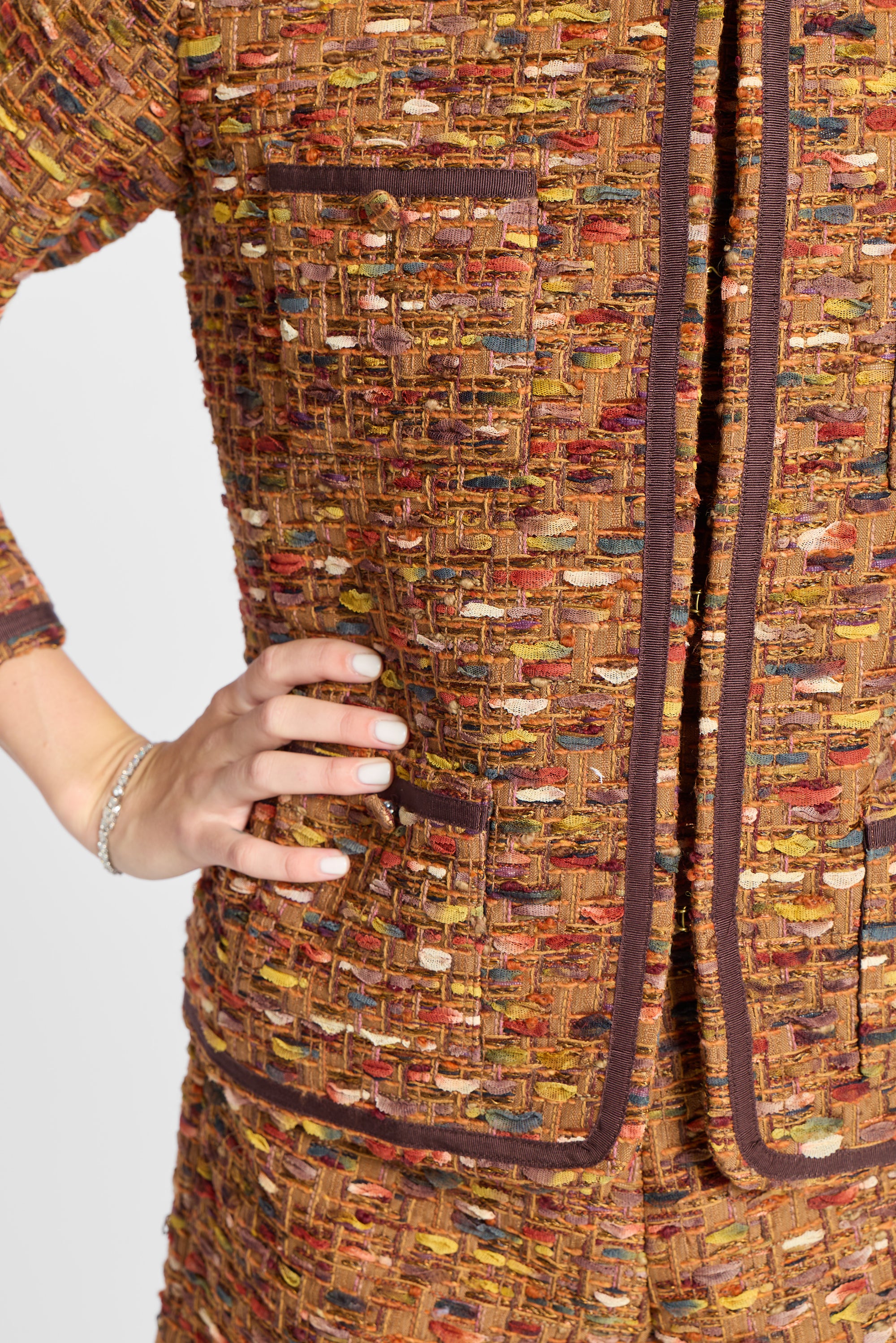 Close-up of a tweed jacket or dress in a multicolor woven blend of brown, gold, red, and cream, with dark brown trim and a hand resting on the hip.