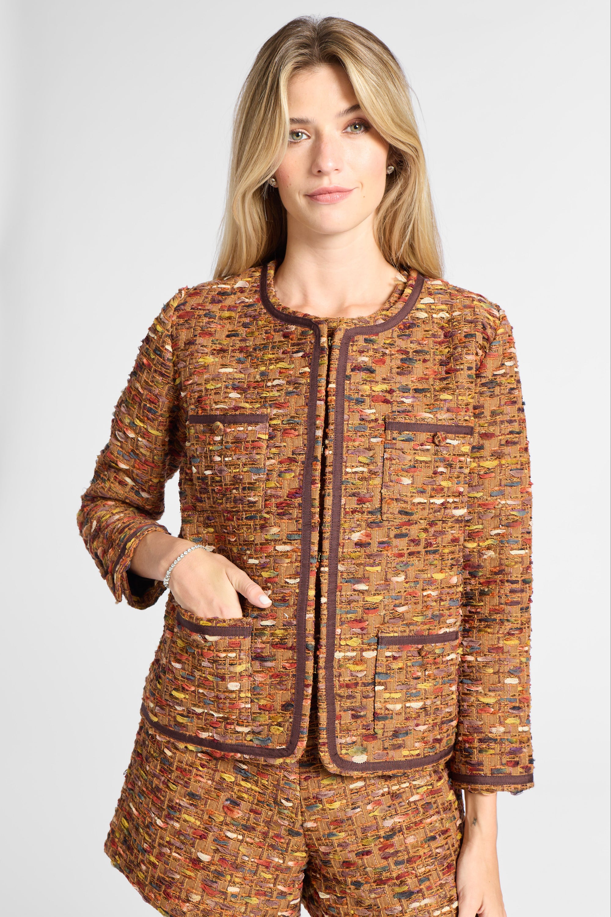 A woman modeling a matching tweed jacket and shorts set in a warm brown, orange, and gold bouclé pattern with dark brown trim, against a light gray studio background.