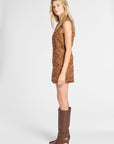 A woman stands in profile wearing a sleeveless, knee-length mini dress with an abstract multicolor pattern in orange, brown, yellow, and red tones, paired with brown knee-high heeled boots, against a light gray studio background.