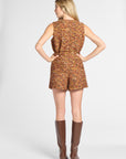 A woman stands with her back to the camera wearing a sleeveless, short romper in a textured multicolor tweed-like fabric with brown and orange tones, paired with tall brown knee-high boots, against a light gray studio background.