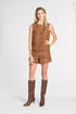 A woman modeling a sleeveless short romper with an abstract multicolor pattern in orange, brown, and yellow tones, paired with knee-high brown boots, against a light gray studio background.