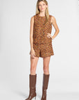 A woman modeling a sleeveless short romper with an abstract multicolor pattern in orange, brown, and yellow tones, paired with knee-high brown boots, against a light gray studio background.