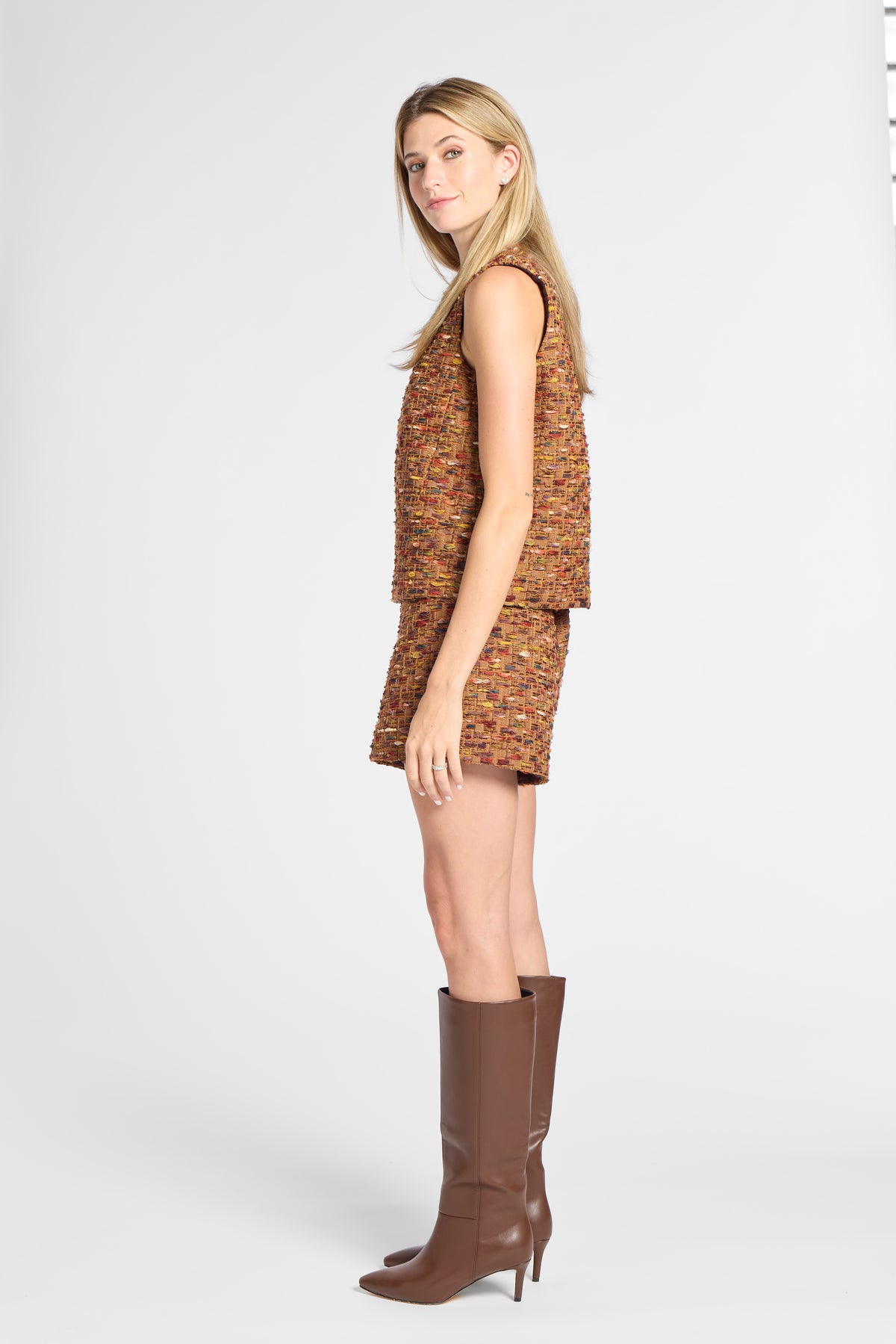 A woman modeling a sleeveless cropped vest and matching shorts set in a textured orange-brown tweed fabric, paired with brown knee-high leather boots, against a light gray studio background.