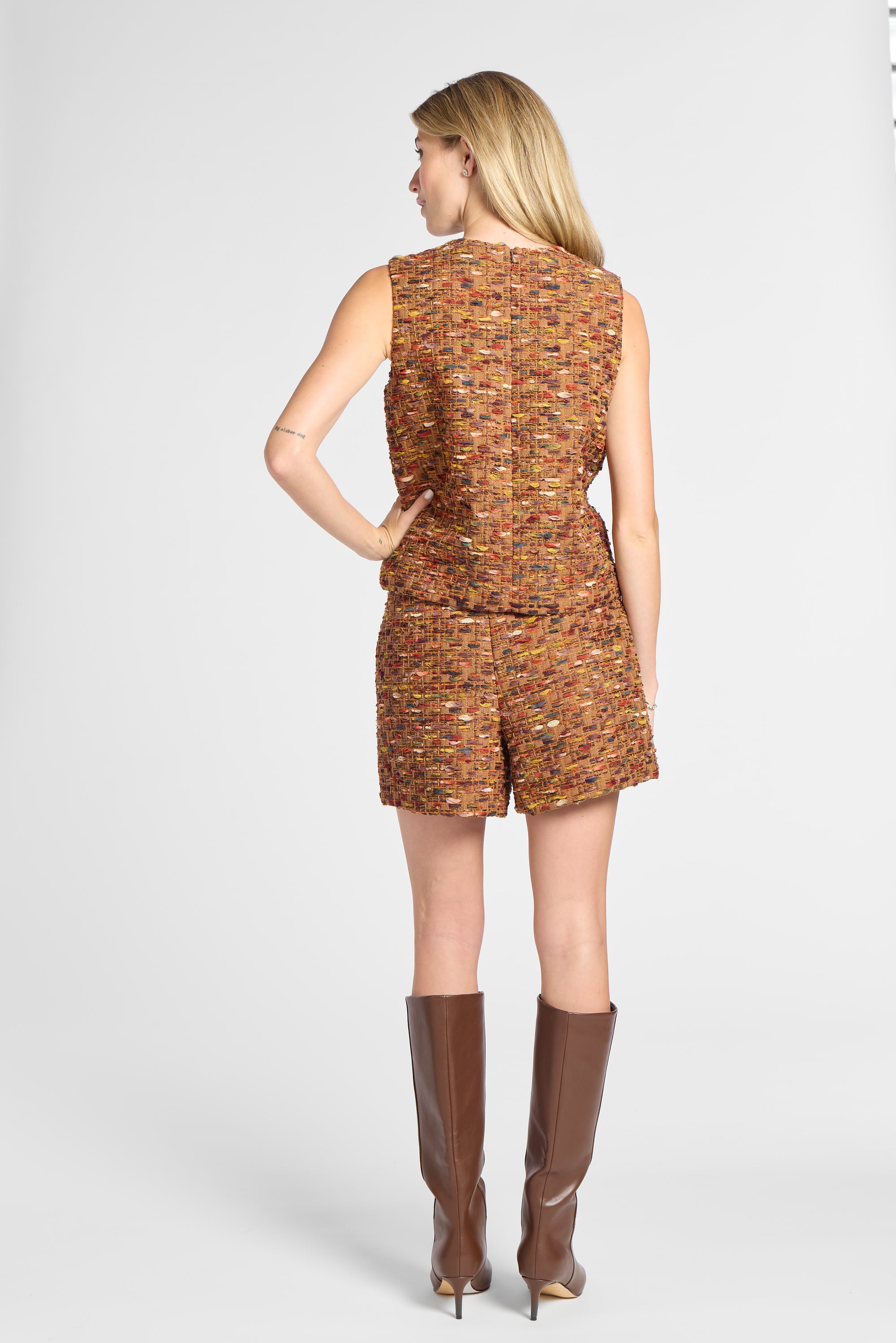 A woman shown from the back wearing a sleeveless, short tweed jumpsuit in a multicolor orange-brown pattern, paired with brown knee-high boots, against a light gray studio background.