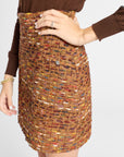 A close-up of a person wearing a brown tweed mini skirt with a small silver button and a brown long-sleeve top, against a light gray background.
