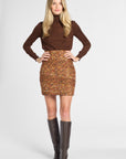A woman modeling a brown turtleneck long-sleeve top and a brown and gold patterned tweed mini skirt, paired with dark brown knee-high leather boots, against a light gray studio background.