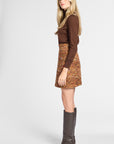 A woman modeling a brown long-sleeve knit top and a brown and orange patterned tweed mini skirt, paired with dark brown knee-high boots, against a light gray studio background.