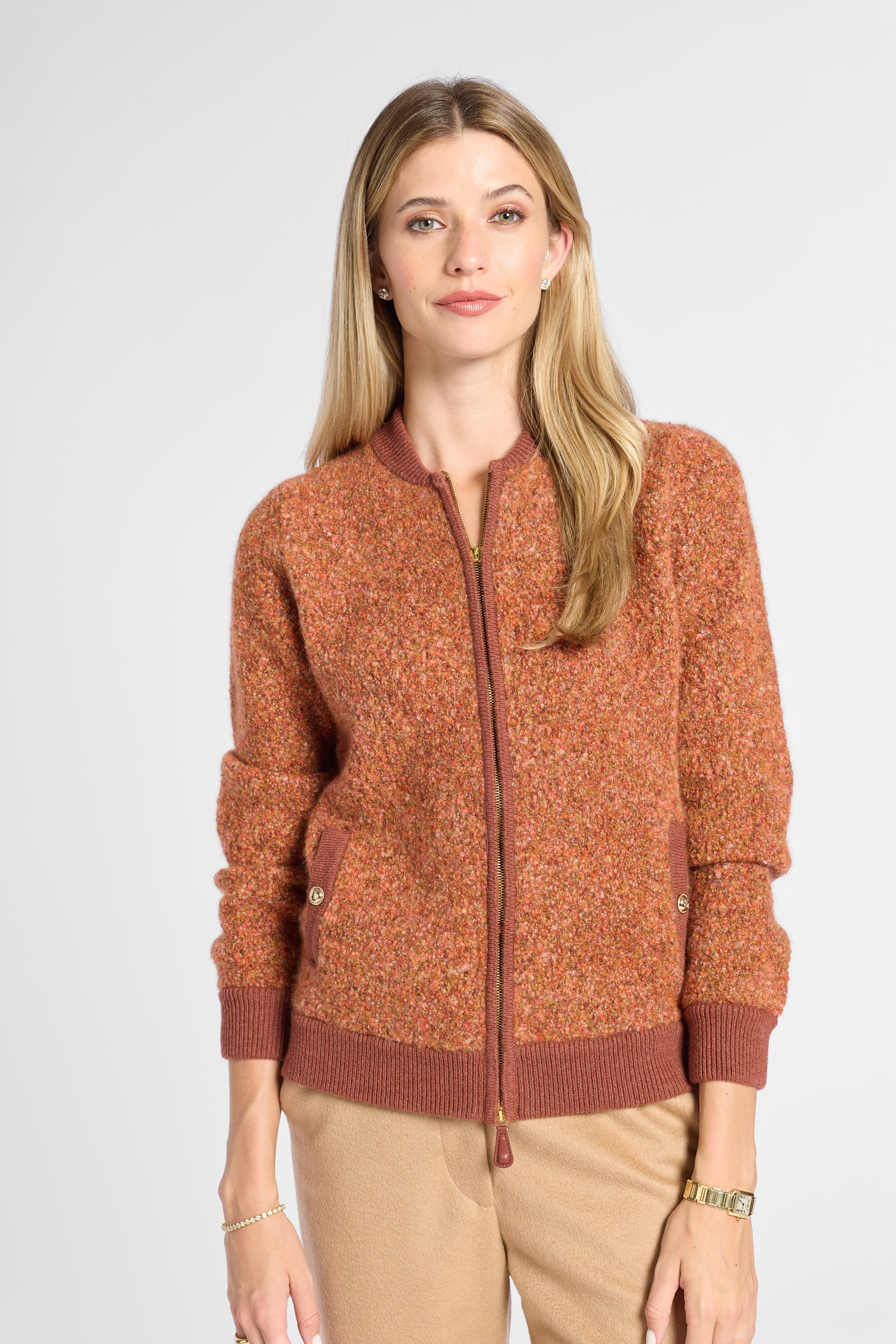 A woman modeling a textured, heathered rust-orange zip-up cardigan with ribbed collar, cuffs, and hem, paired with beige pants, against a light gray studio background.