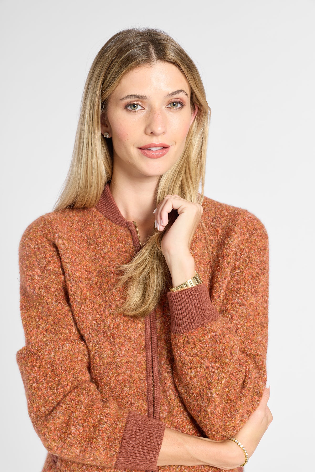 A woman with blonde hair wearing a textured, heathered rust-orange zip-up sweater with ribbed brown collar, cuffs, and hem, posed against a light gray studio background.