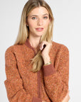 A woman with blonde hair wearing a textured, heathered rust-orange zip-up sweater with ribbed brown collar, cuffs, and hem, posed against a light gray studio background.