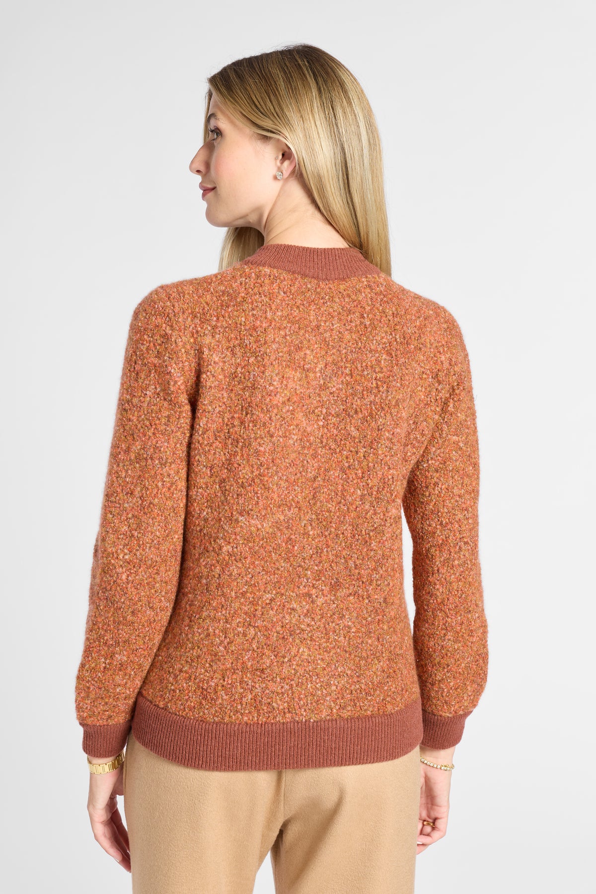 A woman shown from the back wearing a textured, heathered rust-orange knit sweater with solid ribbed trim, paired with beige pants, against a light gray studio background.