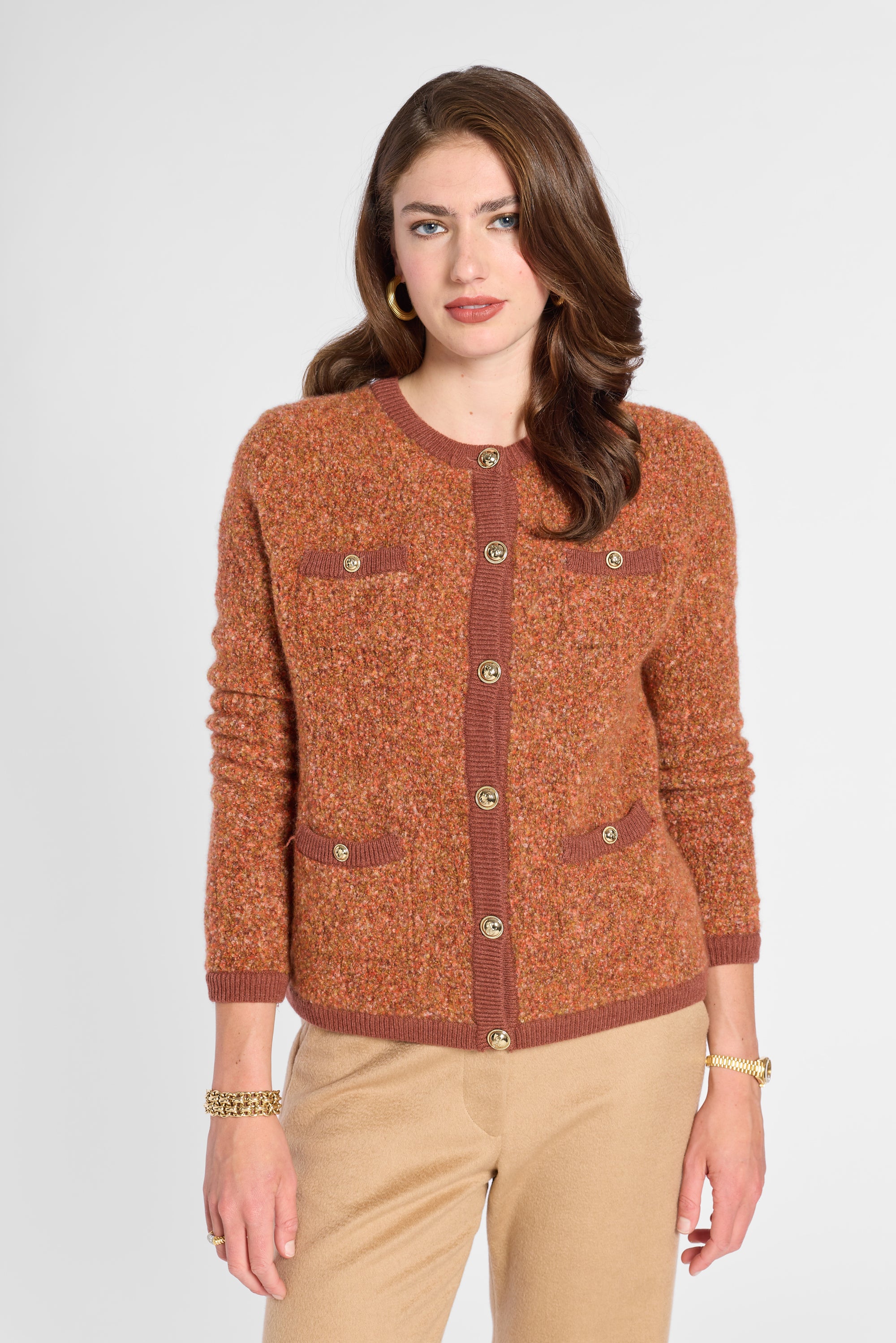 A woman modeling a textured rust-orange cardigan sweater with brown ribbed trim and gold-tone buttons, paired with beige pants, against a light gray studio background.