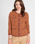 A woman modeling a textured rust-orange cardigan sweater with brown ribbed trim and gold-tone buttons, paired with beige pants, against a light gray studio background.