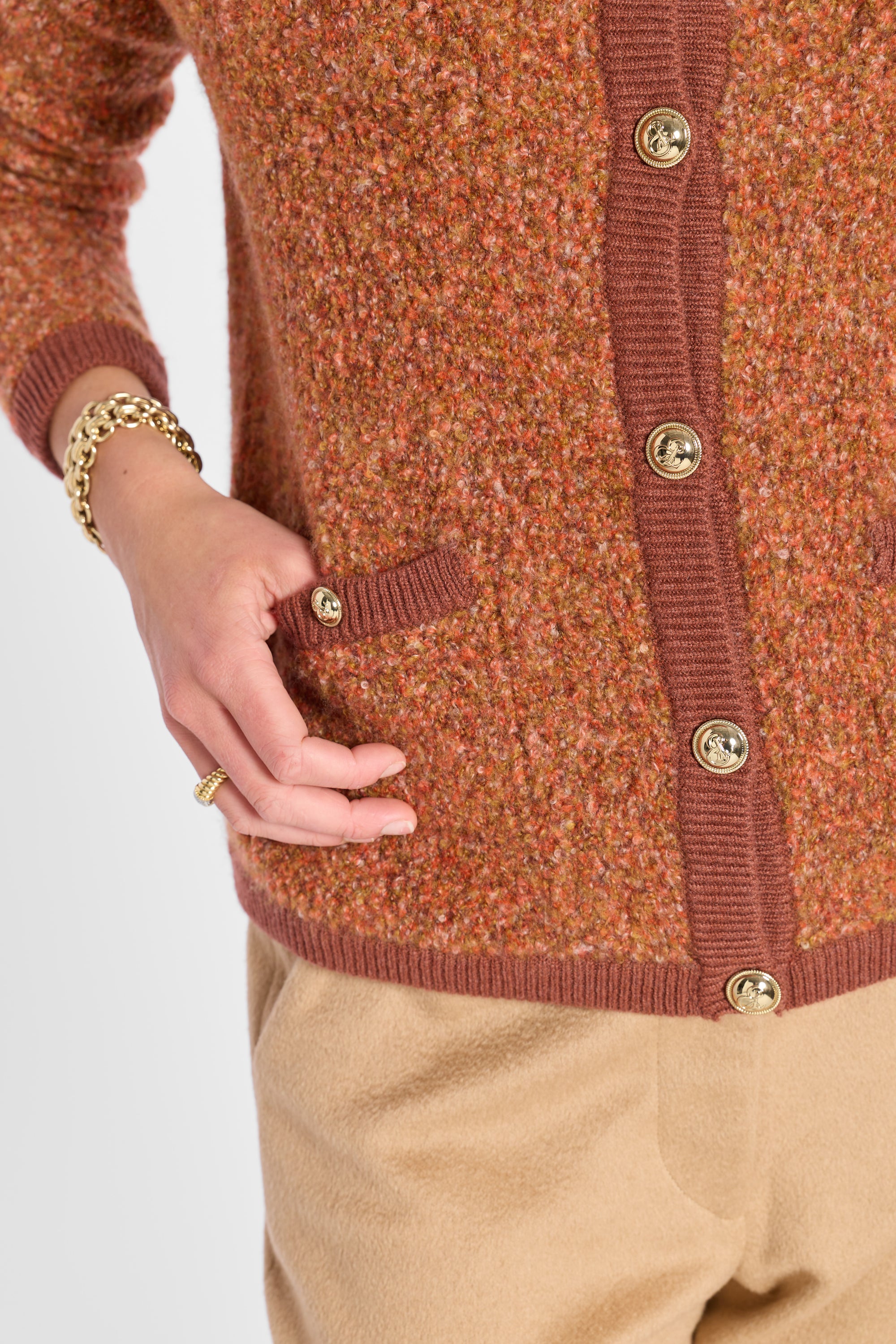 Close-up of a person wearing a rust-orange tweed cardigan with brown ribbed trim and gold-tone buttons, paired with beige trousers. The person's hand is resting on the pocket.
