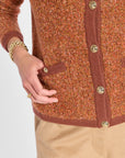 Close-up of a person wearing a rust-orange tweed cardigan with brown ribbed trim and gold-tone buttons, paired with beige trousers. The person's hand is resting on the pocket.