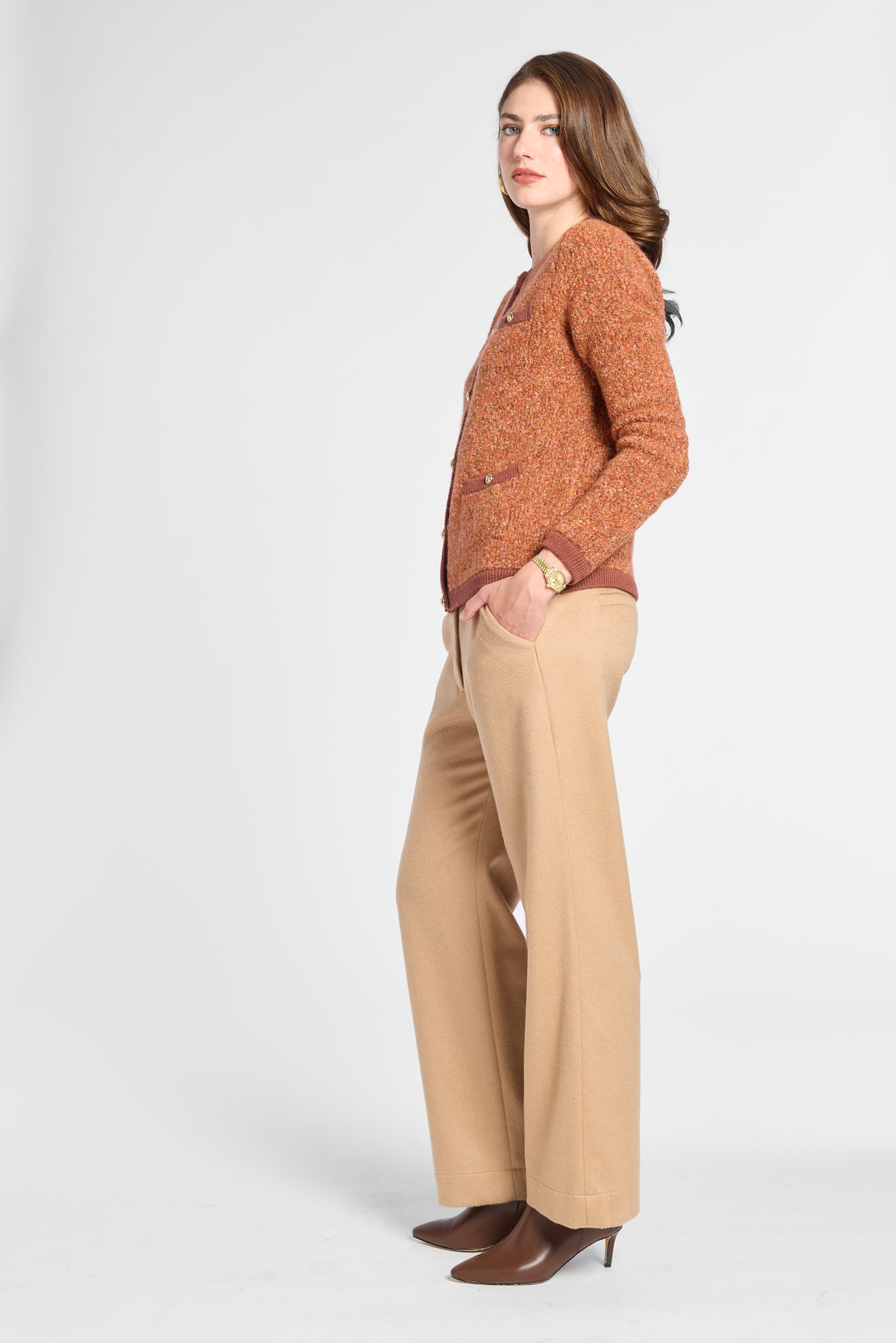 A woman modeling a rust-orange textured knit cardigan sweater, paired with beige wide-leg trousers and brown pointed-toe heels, shown in a side-profile studio shot against a light gray background.