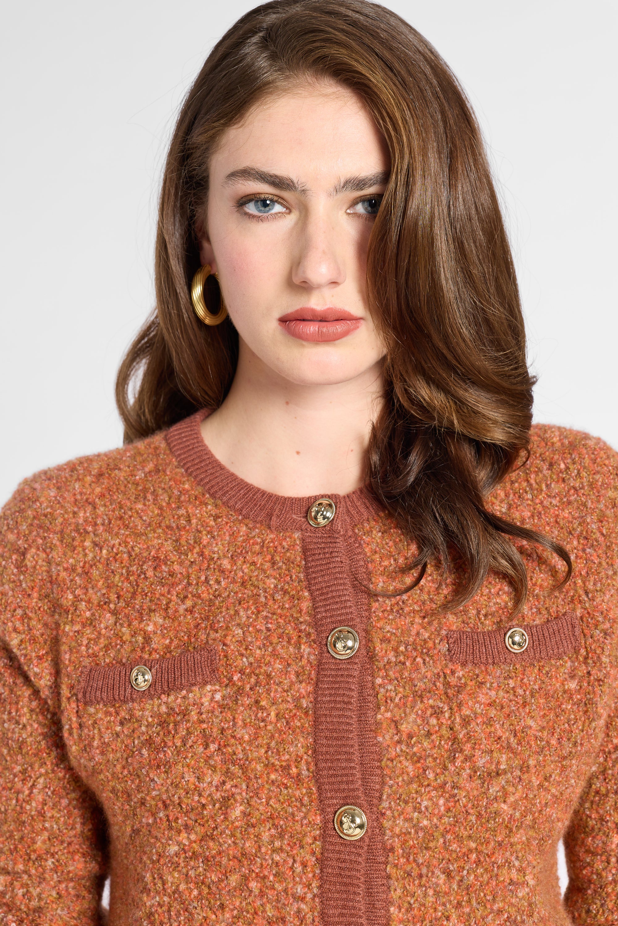 A woman wearing a textured knit cardigan sweater in a warm rust-orange color with brown ribbed trim and gold-tone buttons, shown against a light gray studio background.