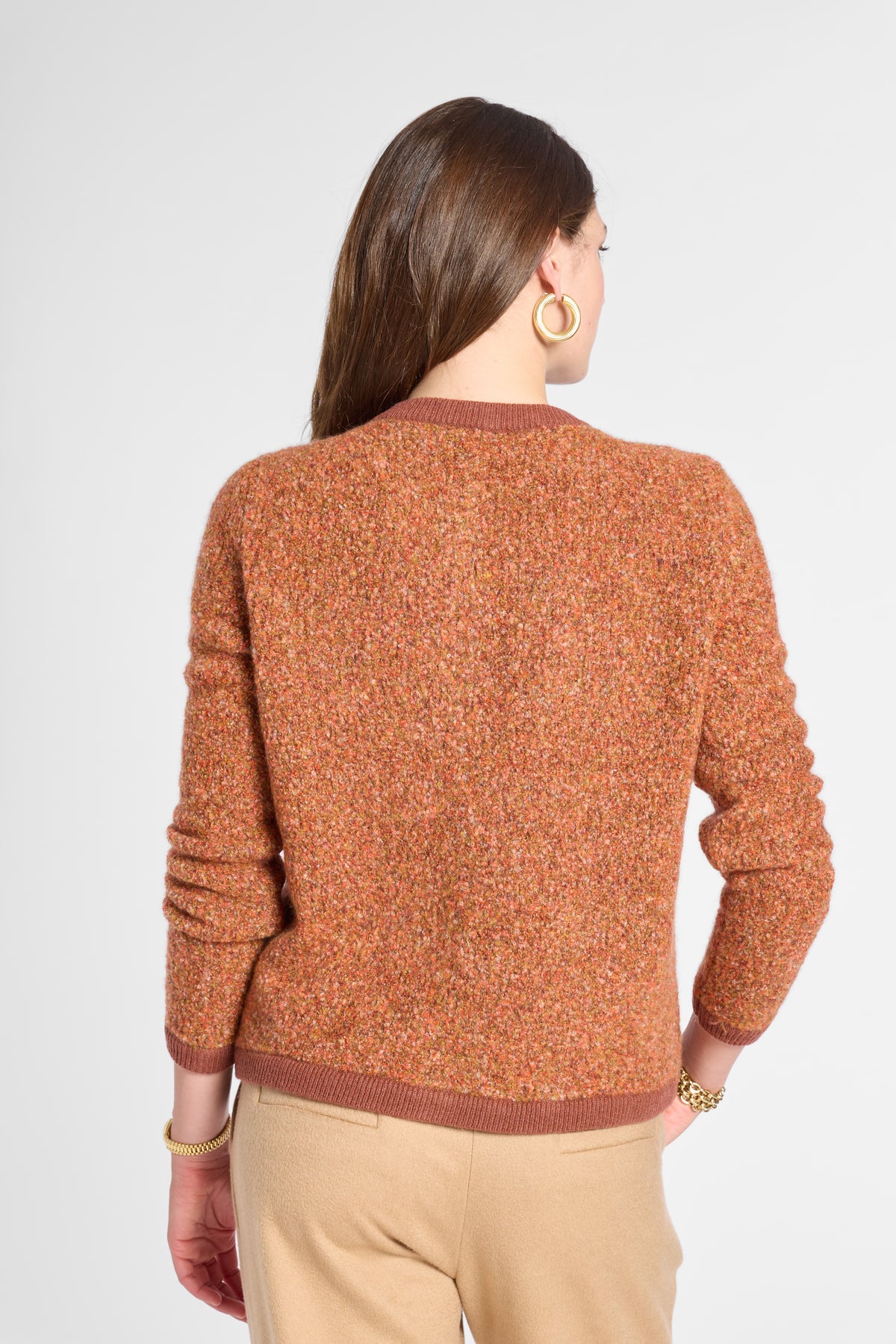 A woman shown from the back wearing a textured, heathered rust-orange knit sweater with brown ribbed trim, paired with beige trousers, against a light gray studio background.