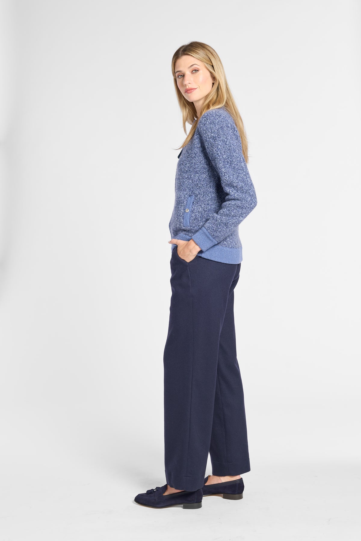 A woman modeling a blue textured knit cardigan sweater and navy wide-leg trousers, standing in profile against a light gray studio background.