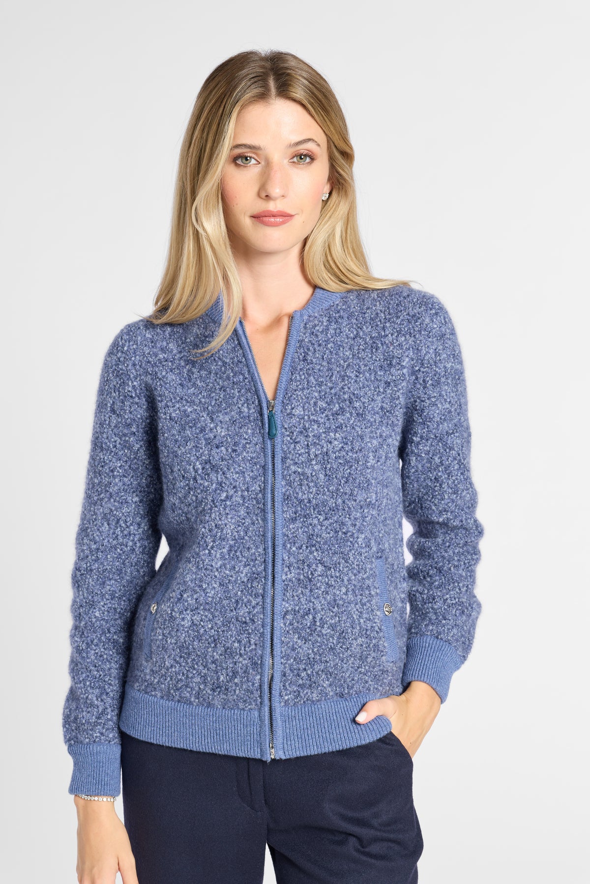 A woman modeling a heathered blue zip-up knit cardigan with ribbed collar, cuffs, and hem, paired with dark navy pants against a light gray studio background.