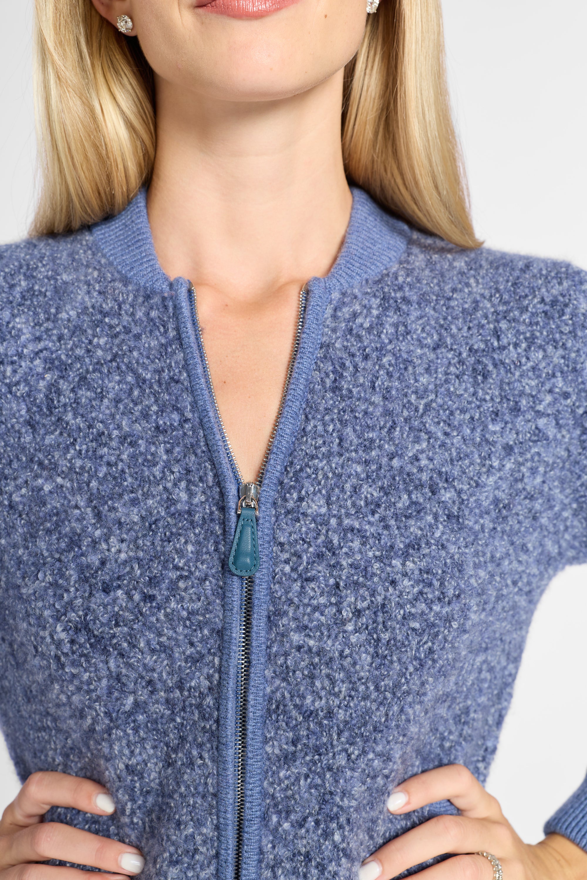 Close-up of a woman wearing a heathered blue zip-front knit sweater with a ribbed collar and cuffs, shown from the shoulders to the hips against a light neutral background.