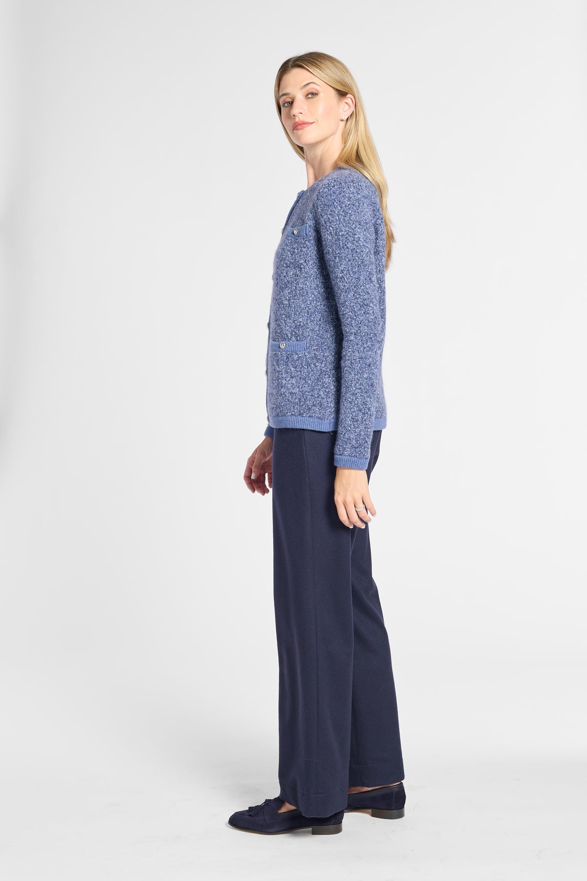 A woman modeling a textured blue knit cardigan sweater paired with navy wide-leg trousers and dark loafers, shown in profile against a plain light gray studio background.