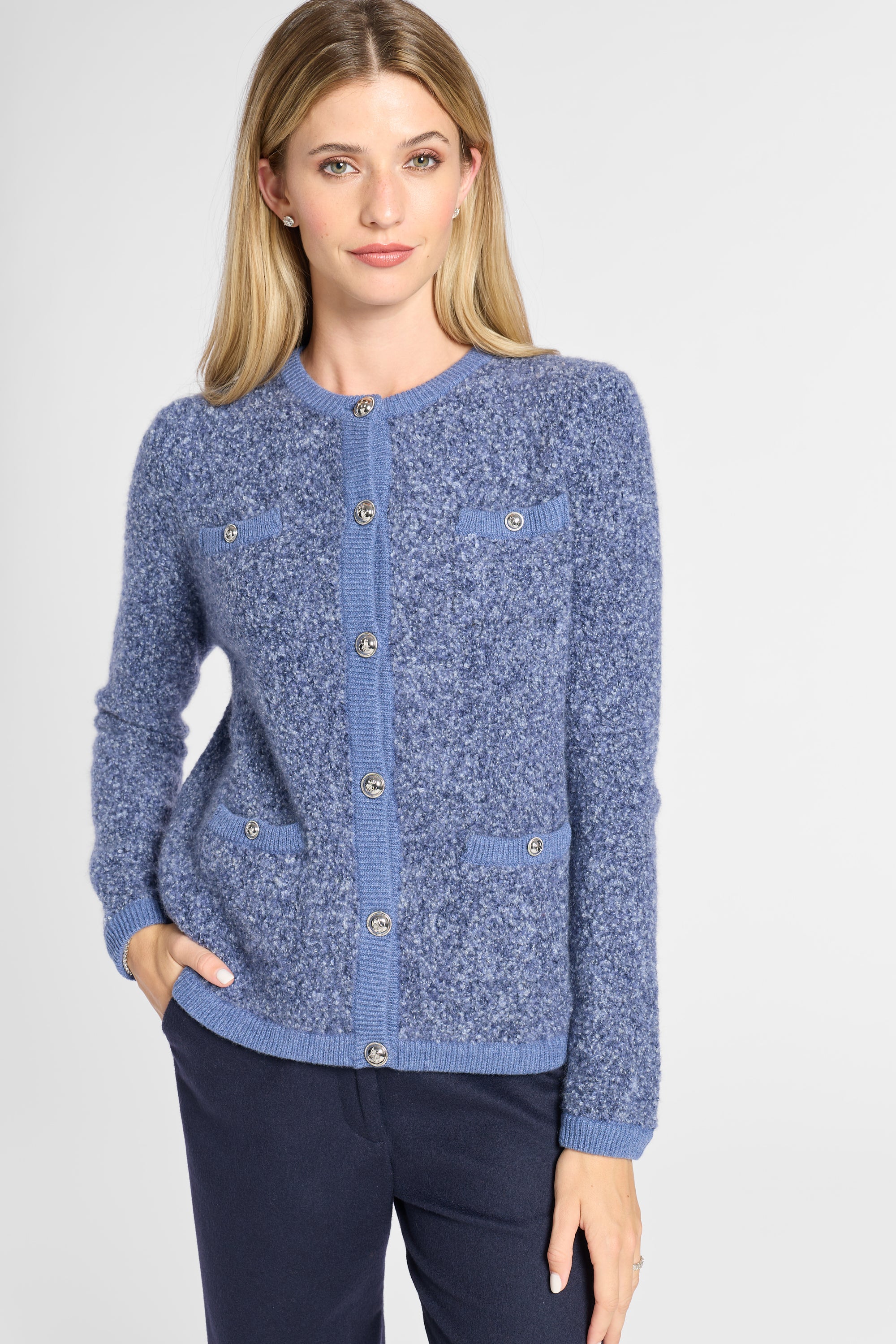 A woman modeling a blue marled knit cardigan sweater with button-front closure and flap pockets, paired with dark navy pants against a light gray studio background.