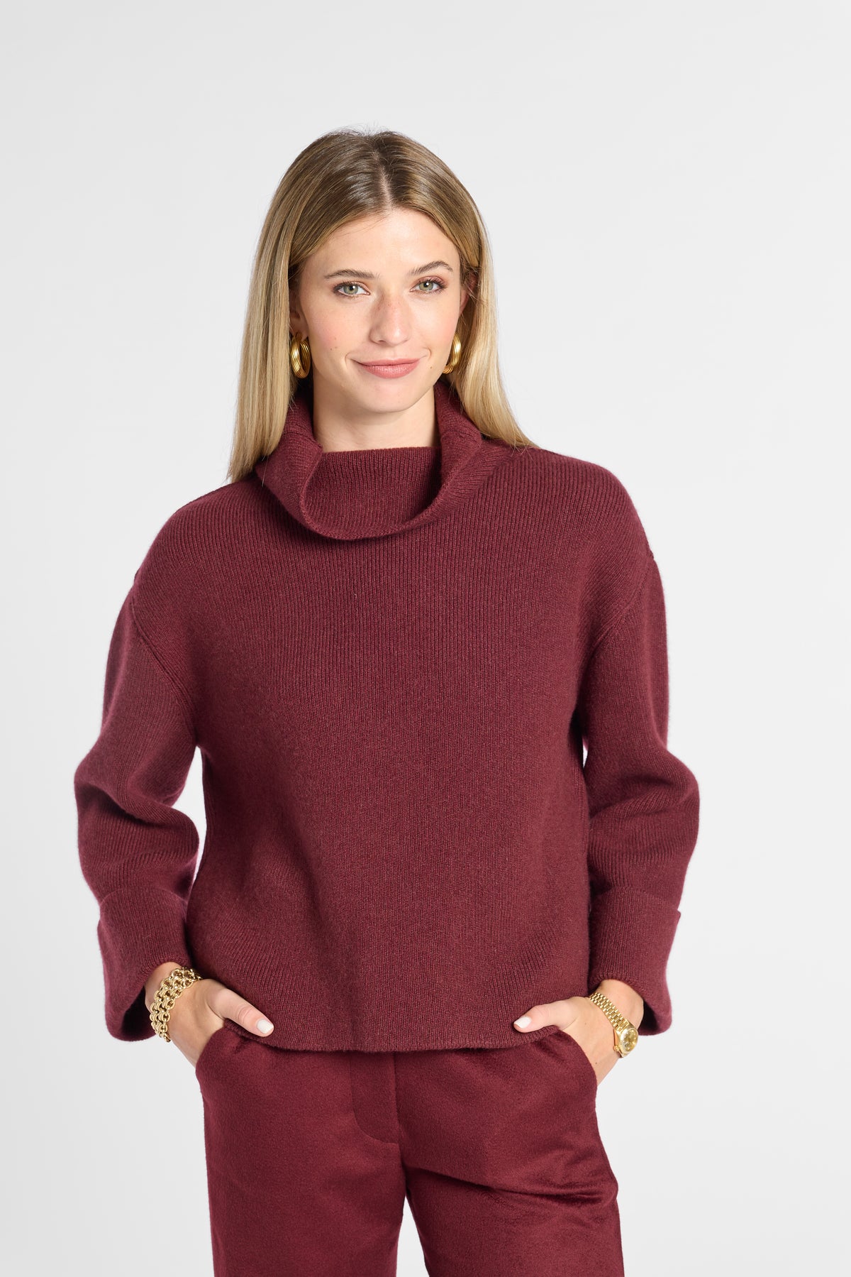 A woman modeling a maroon knit turtleneck sweater and matching trousers against a light gray studio background.